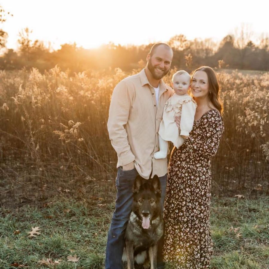 Stryker DeGayner with his wife, their baby daughter and their German shepherd in a field at sunset
